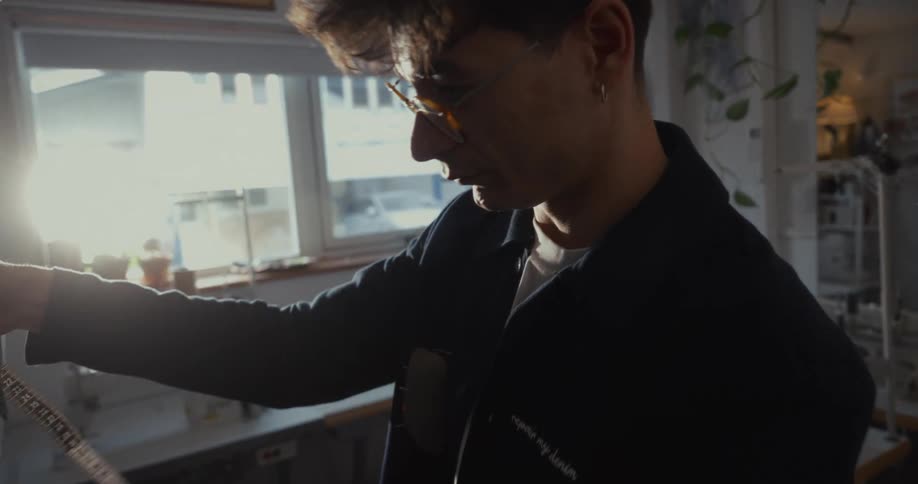 A focused male tailor with glasses meticulously measures a denim jacket sleeve in his sunlit studio