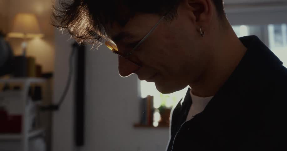 A man with glasses concentrates on his work in a sunlit room, his profile silhouetted by the warm light
