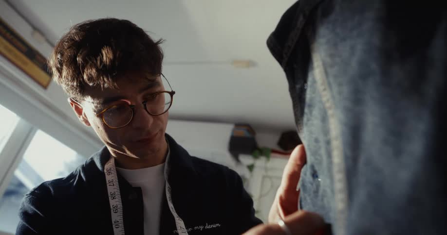 A focused young tailor with glasses carefully pins a denim garment on a mannequin in his sunlit workshop