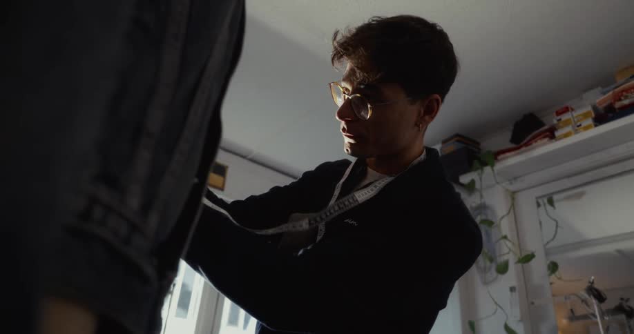 A young male tailor with glasses carefully measures and pins a denim garment hanging in his naturally lit workshop