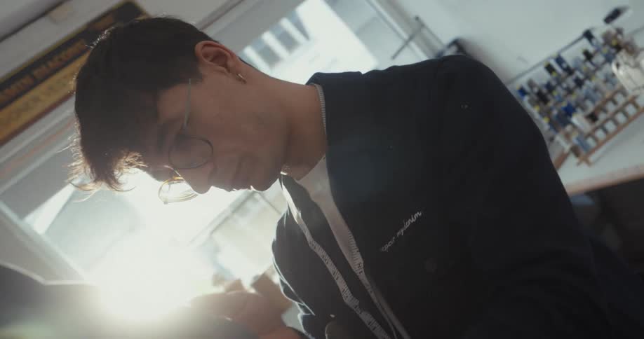 A focused tailor with glasses carefully hand-sews a denim jacket in his sunlit studio