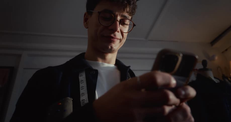 A content young tailor with glasses and a measuring tape smiles while scrolling on his smartphone in his dimly lit workshop