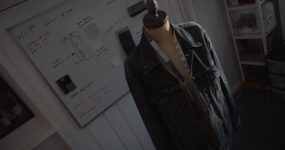 A moody, high-angle shot of a denim jacket on a mannequin in a fashion design workshop with sketches on a whiteboard