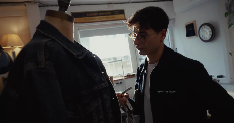 A dedicated young clothing designer meticulously measures a denim jacket on a mannequin in his sunlit workshop