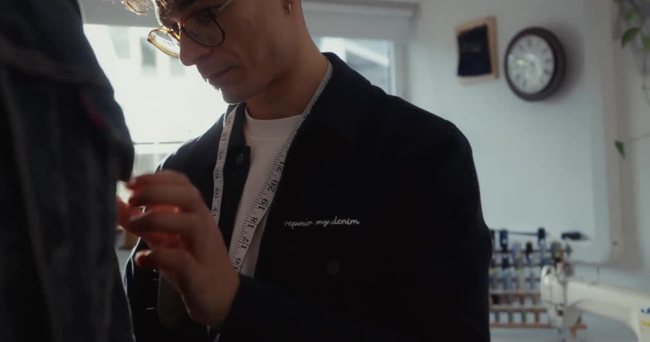 A focused male fashion designer with glasses carefully inspects a denim jacket in his sunlit workshop