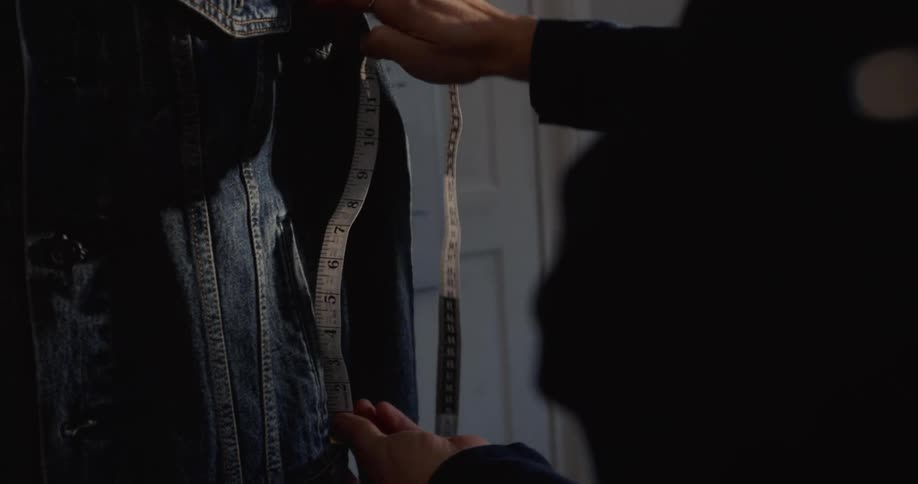Close-up slow motion shot of a seamstress measuring a blue denim jacket with a tape measure in a workshop