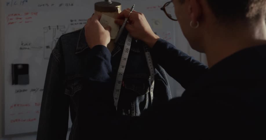 A focused fashion designer takes measurements and notes on a denim jacket fitted on a mannequin in his design studio