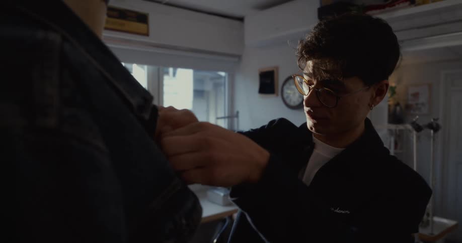 A young tailor with glasses meticulously pins and measures a denim jacket on a mannequin in his sunlit workshop