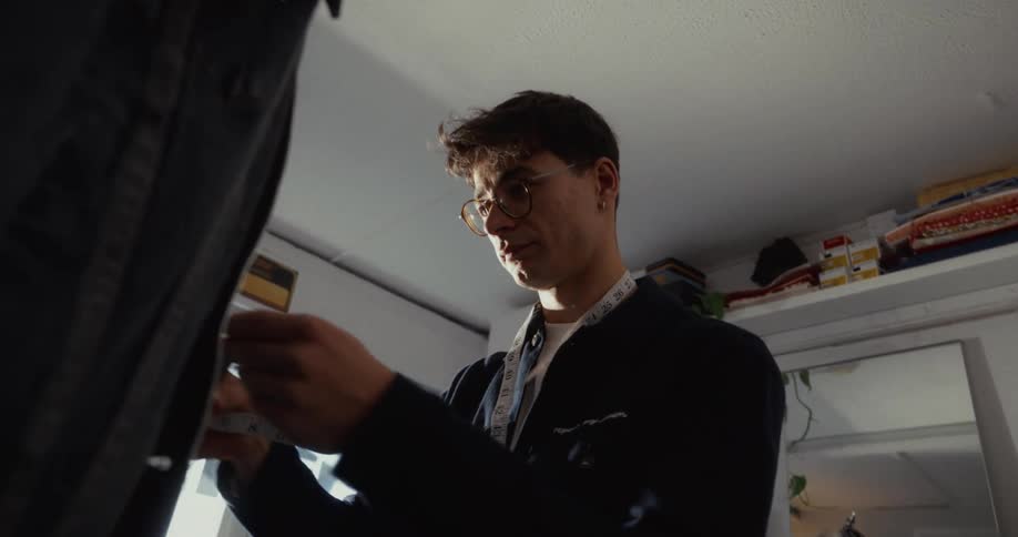 A focused young tailor with glasses carefully measures a denim jacket in his sunlit studio