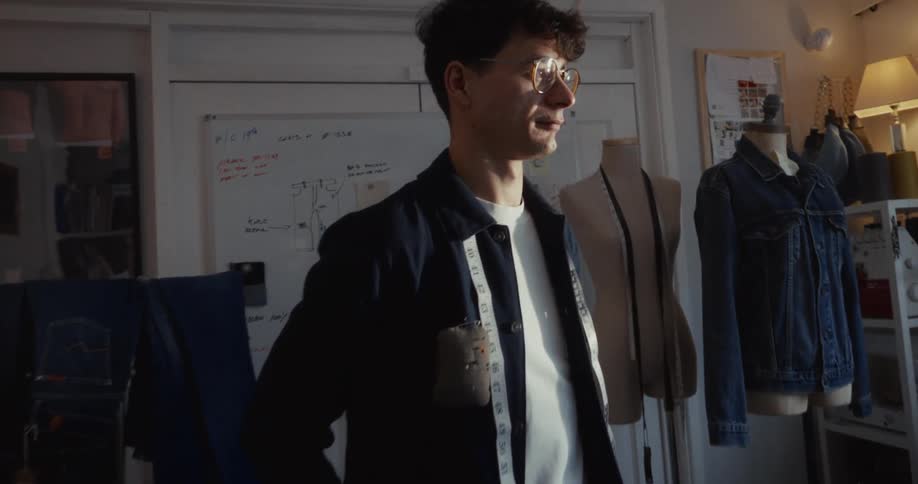 A male fashion designer stands thoughtfully in his sunlit studio, surrounded by his denim creations