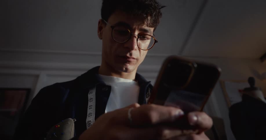 A focused young male fashion designer with glasses and a measuring tape looks at his smartphone in his dimly lit workshop