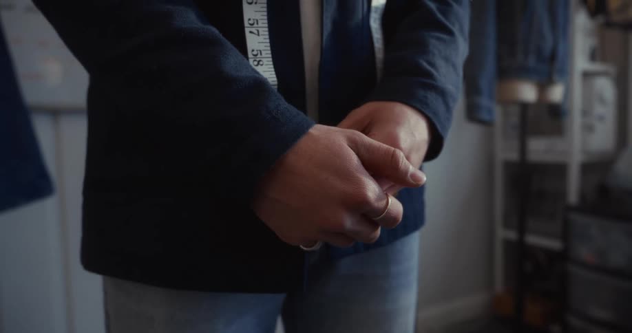 Close-up on the clasped hands of a tailor with a measuring tape draped around their neck in a design studio