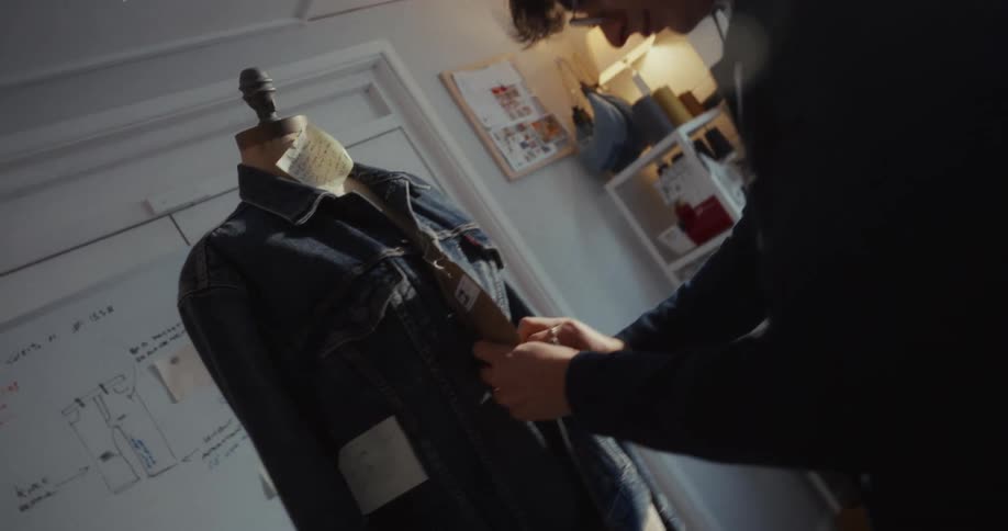 A young male fashion designer meticulously pins a tag on a denim jacket displayed on a mannequin in his creative workshop