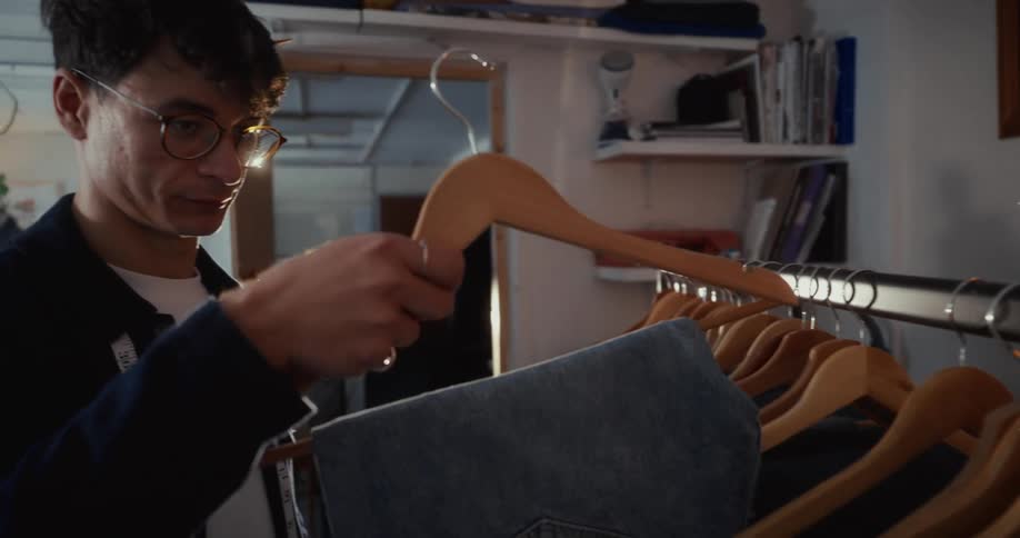 A male tailor wearing glasses carefully inspects a pair of denim jeans on a clothing rack in his sunlit workshop