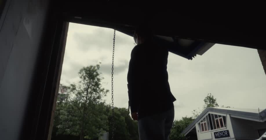A silhouetted person opens a large industrial garage door with a chain, revealing an overcast day outside