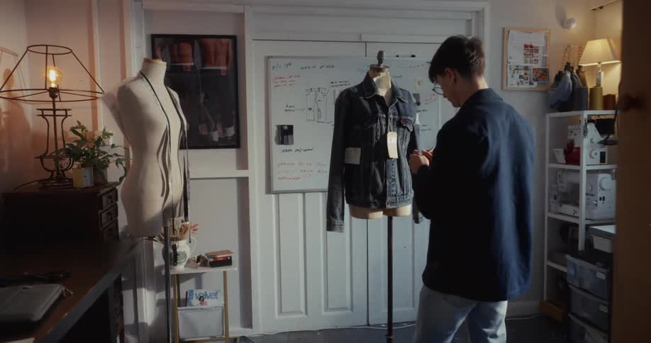 Slow push-in shot reveals a young male fashion designer meticulously working on a denim jacket on a mannequin in his studio