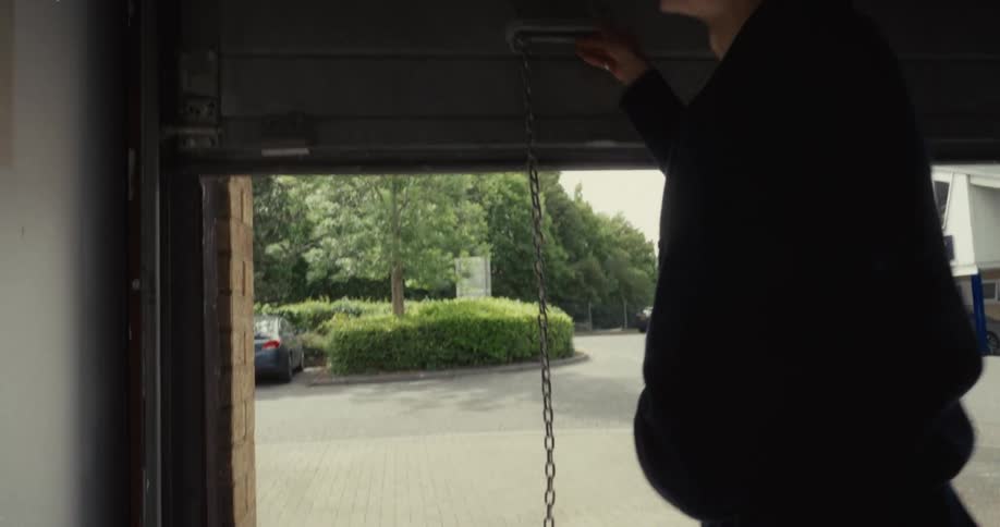 A man opens a metal roll-up garage door from the inside, letting daylight flood into the dark space