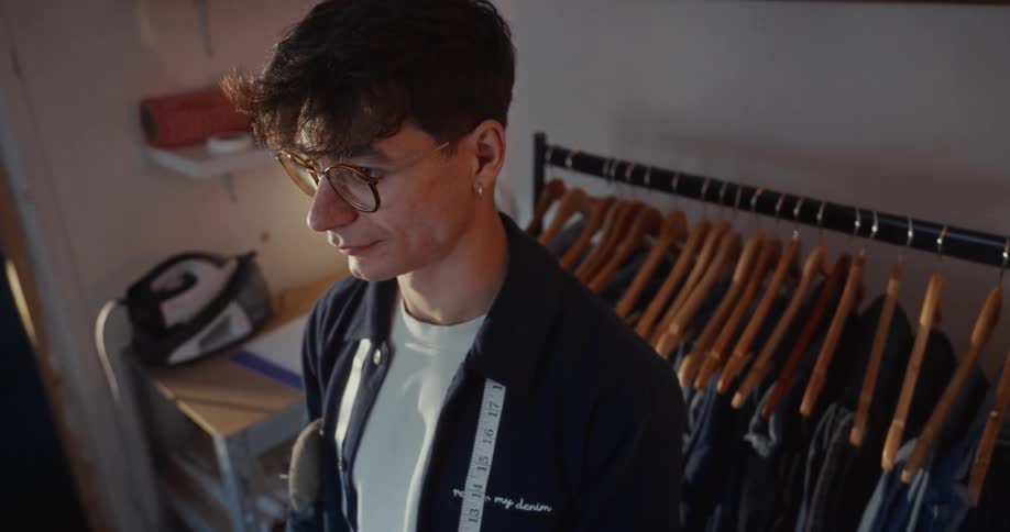 A young tailor with glasses and a measuring tape around his neck works thoughtfully in his cozy studio
