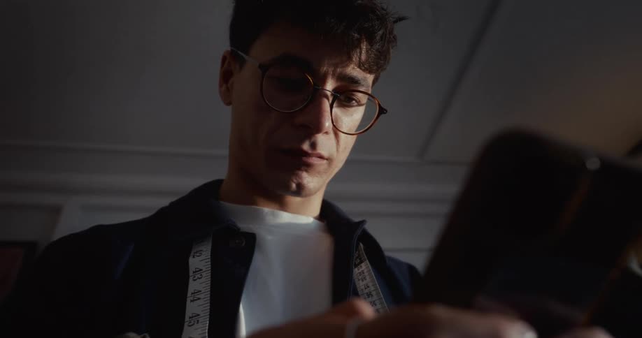 Low-angle close-up of a focused male tailor in glasses scrolling on his smartphone in a dimly lit workshop