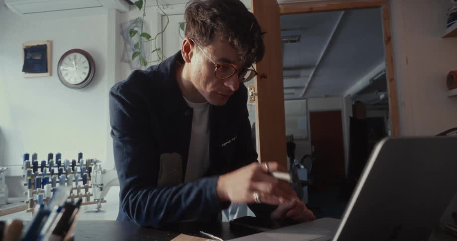 A focused young designer with glasses works on his laptop and smartphone in a creative workshop