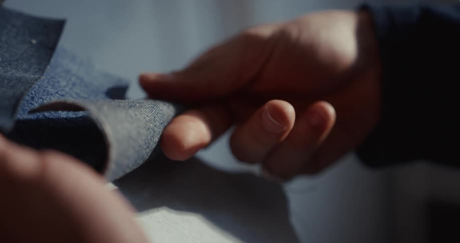 A designer's hands carefully inspect and feel different shades of denim fabric swatches in slow motion
