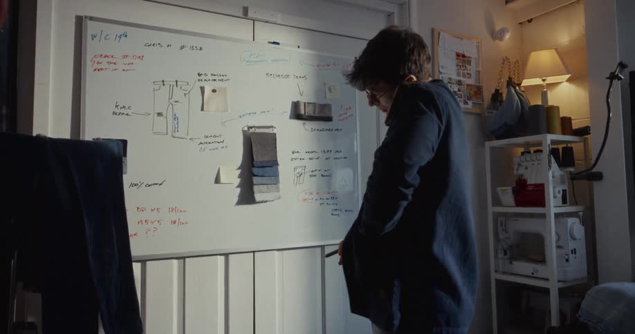 Focused fashion designer talks on the phone while reviewing denim sketches and fabric swatches on a whiteboard in his studio