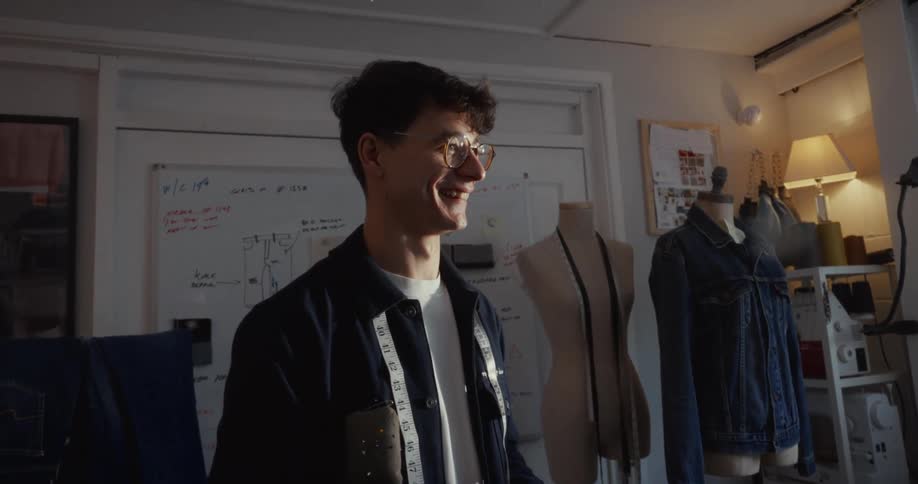 A smiling young tailor stands proudly in his sunlit design studio surrounded by his denim clothing creations