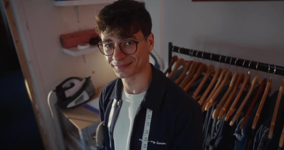 A proud young male tailor with glasses smiles warmly at the camera in his sunlit workshop