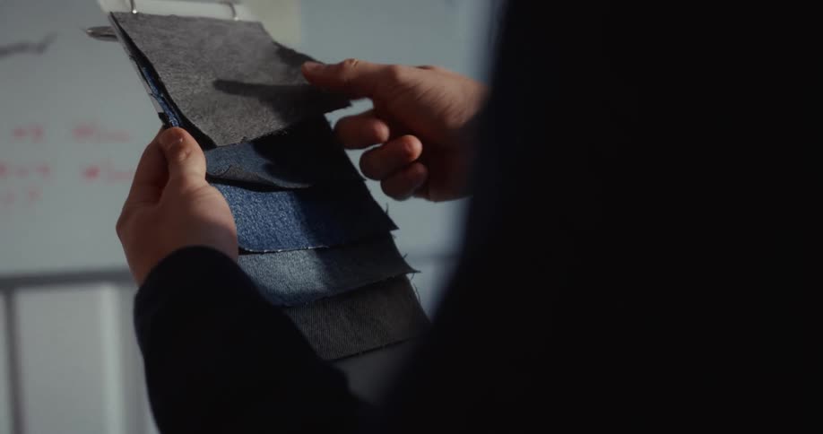 A designer's hands carefully flip through a book of various denim fabric swatches in a sunlit studio