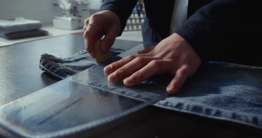 A tailor's hands carefully measure and mark a line on a pair of blue jeans with a ruler and chalk in a workshop