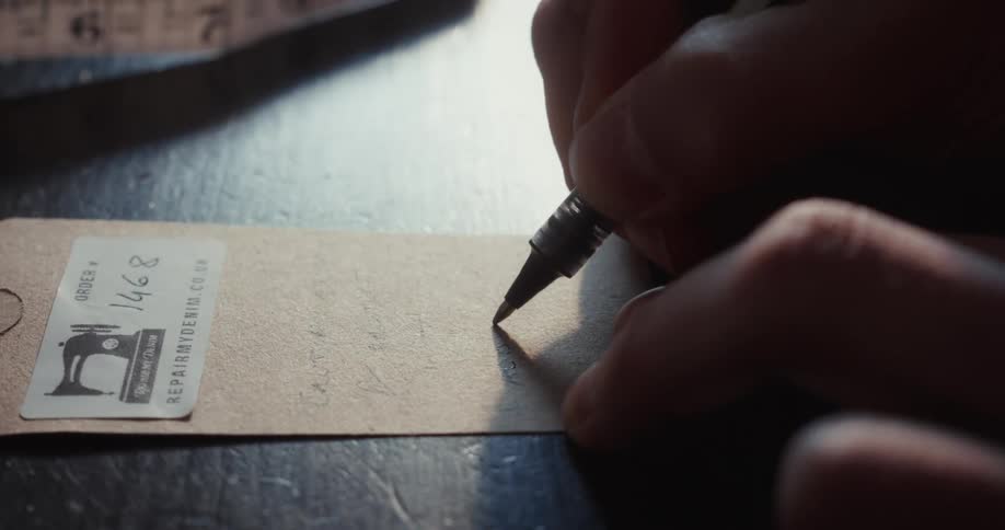 A tailor carefully writes down customer order notes on a brown paper slip in a sunlit workshop
