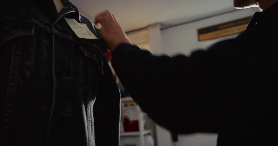 A smiling male tailor pins a handwritten note onto a classic denim jacket on a mannequin in his sunlit workshop