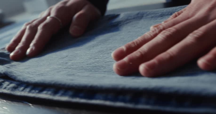 A person's hands gently smooth the fabric of a pair of blue jeans in a slow-motion close-up shot