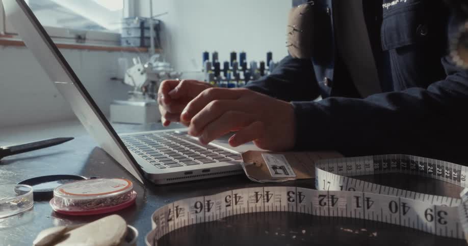A clothing designer works on their laptop, surrounded by the tools of their trade in a sunlit studio