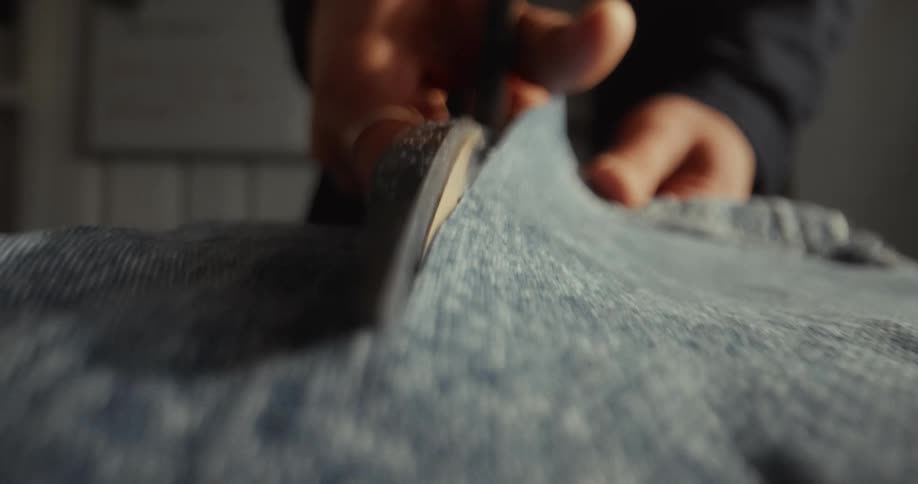 A close-up shot of hands using sharp scissors to cut through blue denim fabric in slow motion