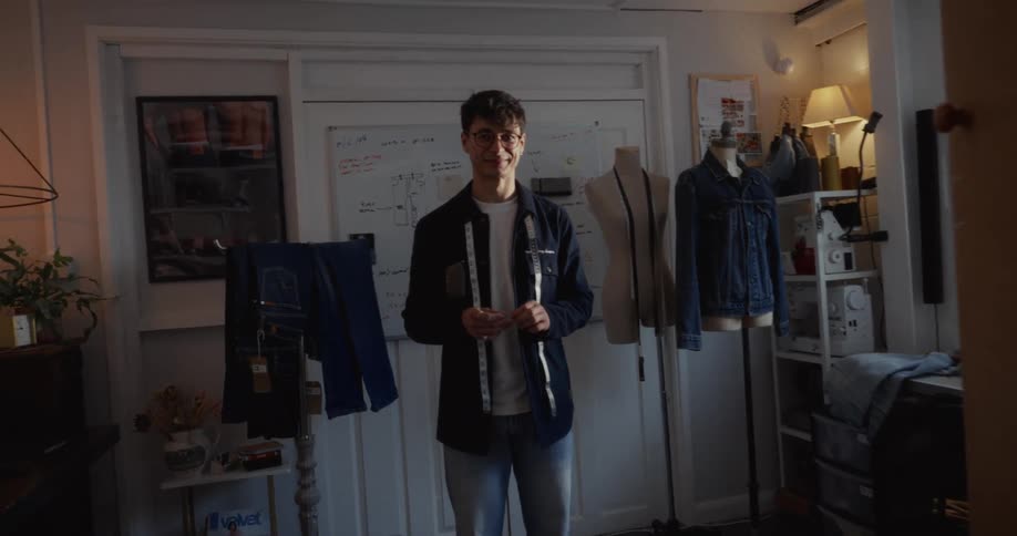 A slow dolly-in shot reveals a confident young fashion designer smiling in his creative workshop surrounded by his denim creations