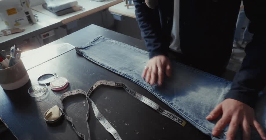A tailor in a workshop carefully folds and smooths a pair of blue jeans on a dark table in slow motion
