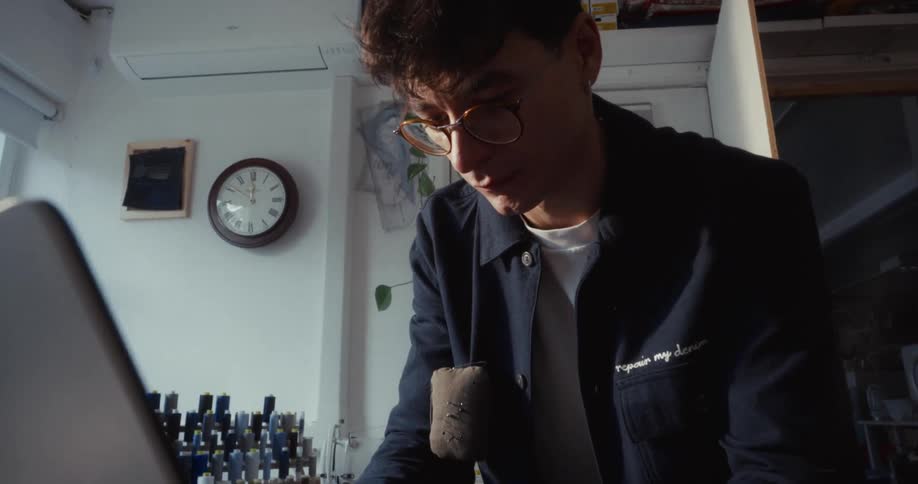 A focused young fashion designer wearing glasses works on his laptop at a desk in his sewing workshop