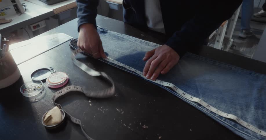A tailor's hands carefully measure the inseam of a pair of blue jeans laid out on a workbench in a workshop