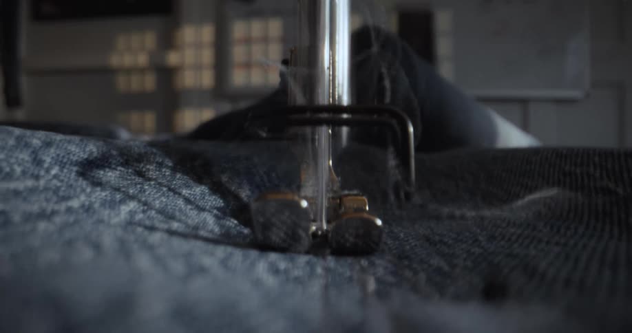 A detailed macro close-up of a sewing machine needle stitching a seam on blue denim fabric