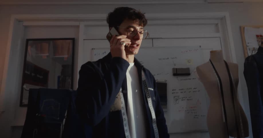 A young male fashion designer with glasses and a measuring tape around his neck talks on the phone in his studio