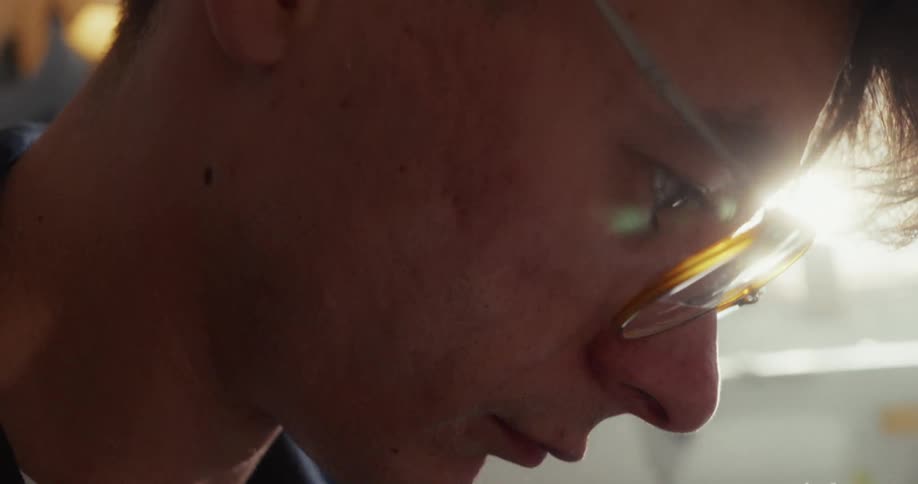 A young man with glasses concentrates intently, his face illuminated by a bright backlight in a close-up profile shot