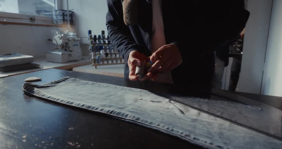 A close-up shot of a tailor's hands carefully selecting the perfect thread to mend a hole in a pair of denim jeans in a workshop