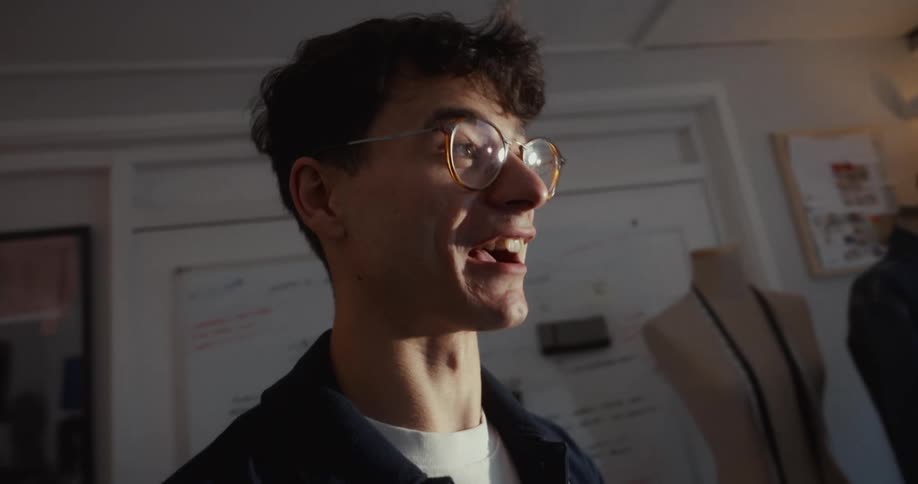 An enthusiastic young male fashion designer with glasses speaks passionately while standing in his sunlit creative studio