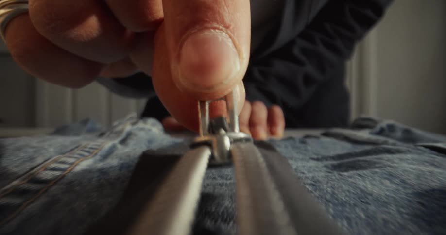 An extreme close-up shot of a hand slowly zipping up the metal zipper on a piece of blue denim clothing