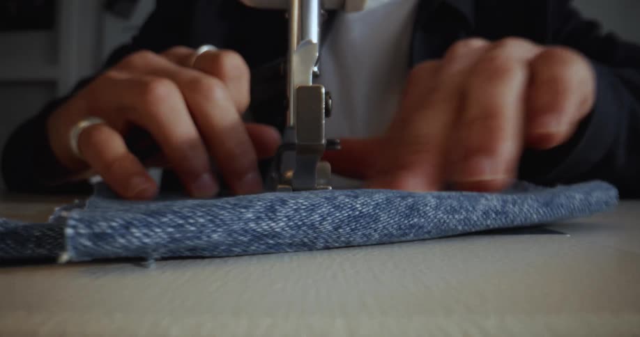 A detailed, front-facing shot shows hands skillfully guiding blue denim fabric through a sewing machine to create a seam