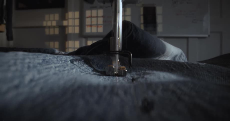 A sewing machine's needle and presser foot move over blue denim fabric in an atmospheric, close-up shot