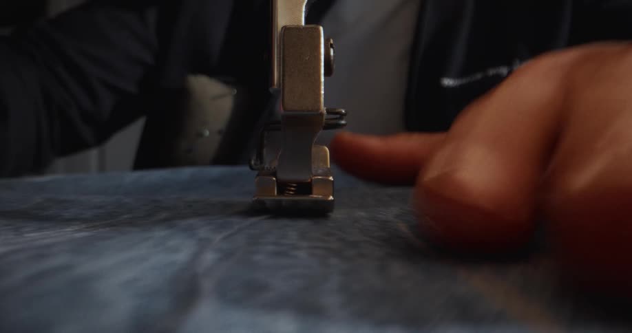 An extreme close-up shot captures the presser foot of a sewing machine stitching a straight seam on blue denim fabric
