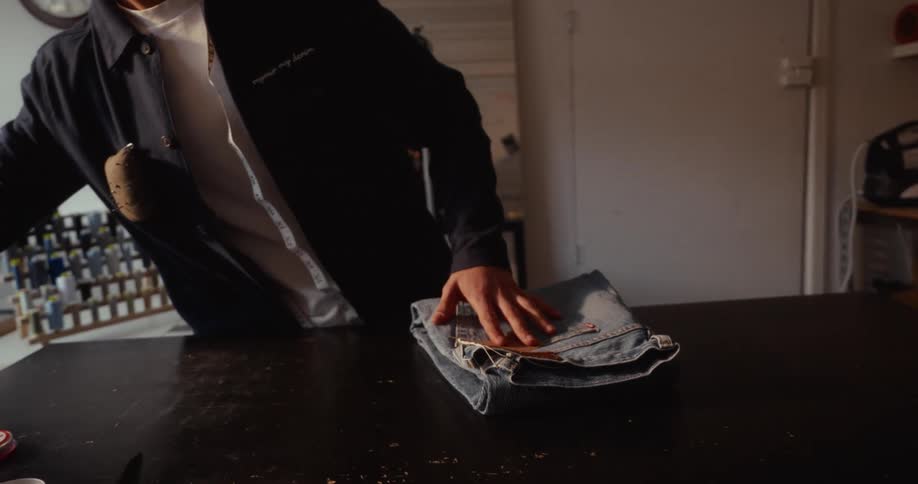 A clothing designer in a studio neatly folds a pair of blue jeans and packs them into a paper bag