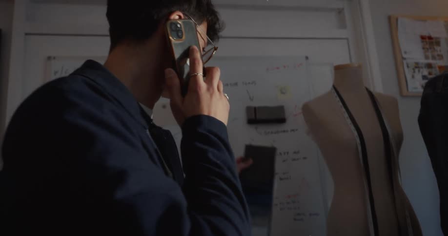 A young male fashion designer talks on the phone while examining fabric swatches and design notes on a whiteboard in his sunlit studio